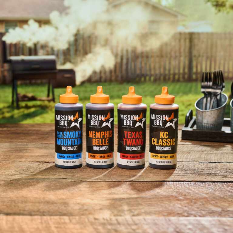 Four bottles of BBQ sauce on a table in a backyard with a smoker inthe background
