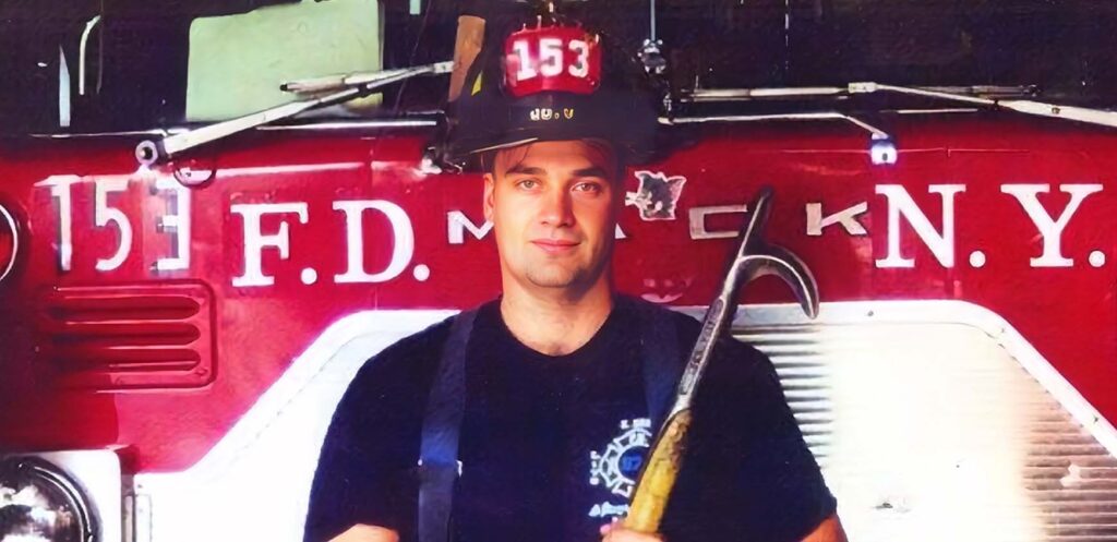 FDNY firefighter Stephen Siller in his helmet, holding a firefighting tool in front of a red fire truck.